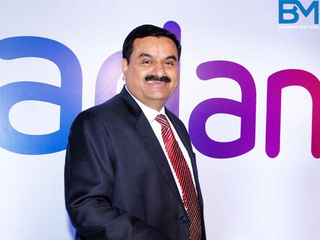 Gautam Adani, CEO of Adani Enterprises standing in fornt of Adani logo on background of stage smiling